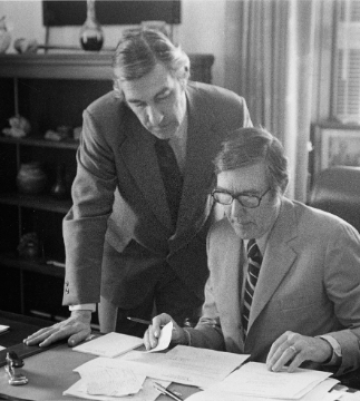 Two men in suits in an office looking at documents, one seated, and the other looking over his shoulder.