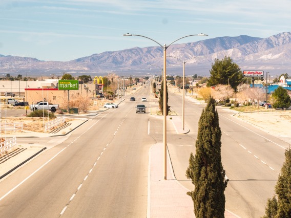 A photo of a paved desert street with a few cars, sparse trees, and mountains in the background against a pale blue sky.