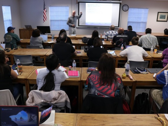 A view of a full classroom from the back row with Dr. Stephen Cornell gesturing near a projector screen at the front of the room