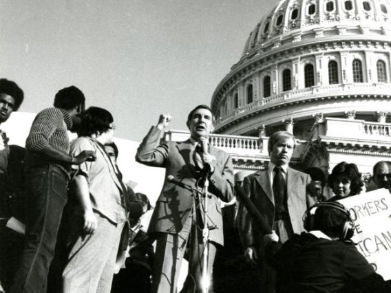 Morris K. Udall on the Capital steps behind a microphone with one first raised in the air. A crowd around him listens as he speaks.