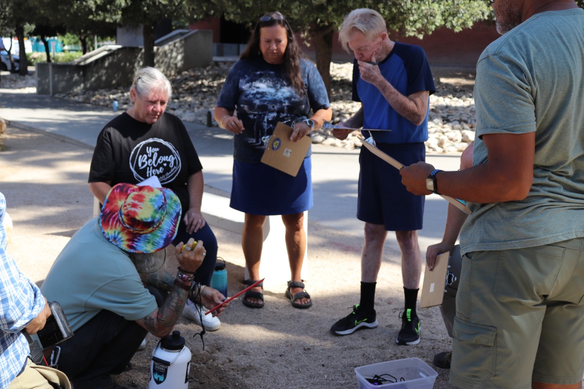 A man in a tie-dye bucket hat kneels over a whole dug in the dirt as a circle of teachers with clipboards look on.