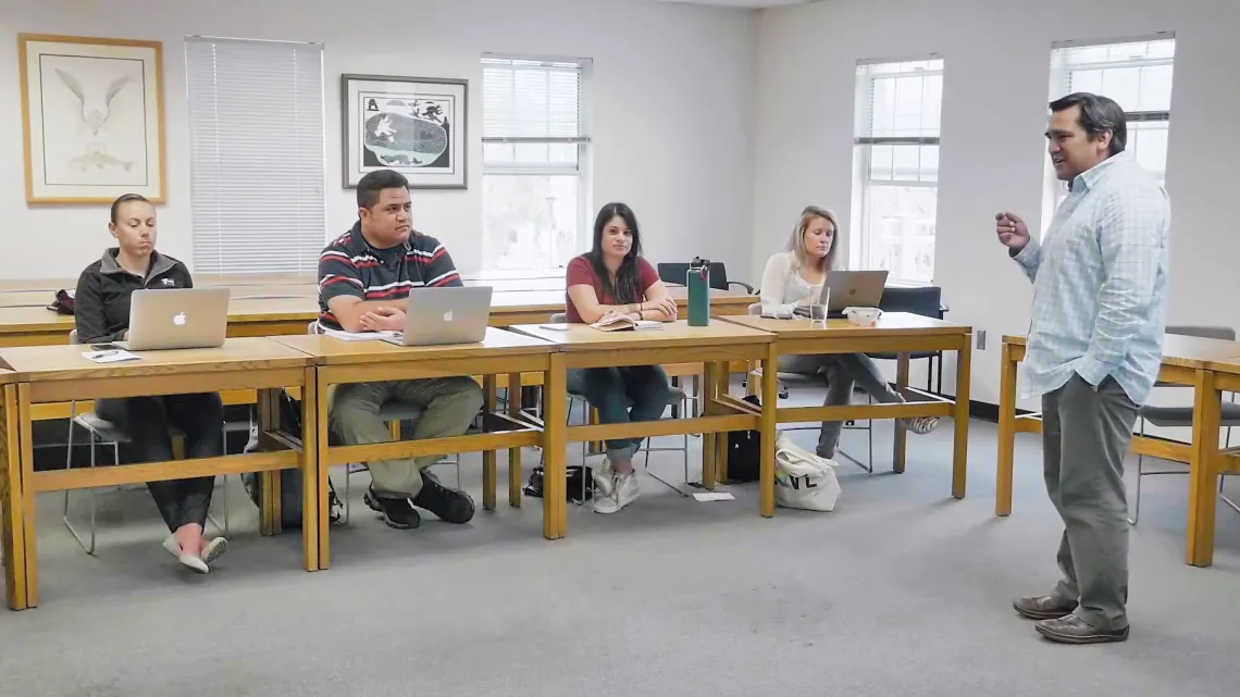 Tory Fodder stands to the right of the image in a blue and white checkered button-up shirt. Toward the left are four students sitting in a single row at yellow-hued wooden desks. Three of the four have silver laptops open in front of them.