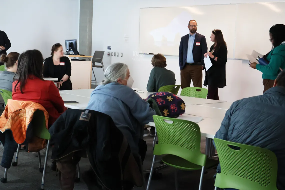 Ladd Keith and Leslie Ethen stand at the front of a classroom with several workshop participants seated in front of them at white tables in green folding chairs.