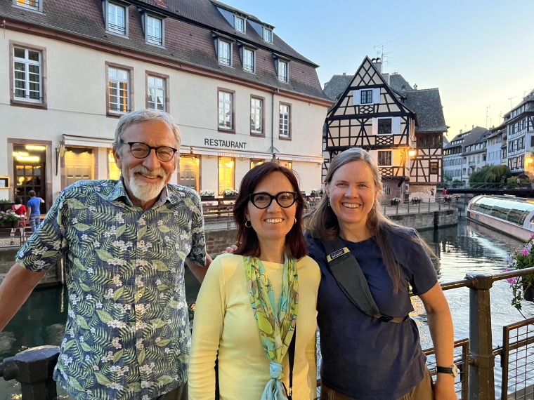 From left to right, Larry Fisher (floral shirt, black-rimmed glasses, goatee), Adriana Zuniga-Teran (yellow shirt, blue and yellow scarf, black-rimmed glasses), and Larry's wife Tahnee Robertson pose for a photo by a canal in Strasborg, France in July 2023.