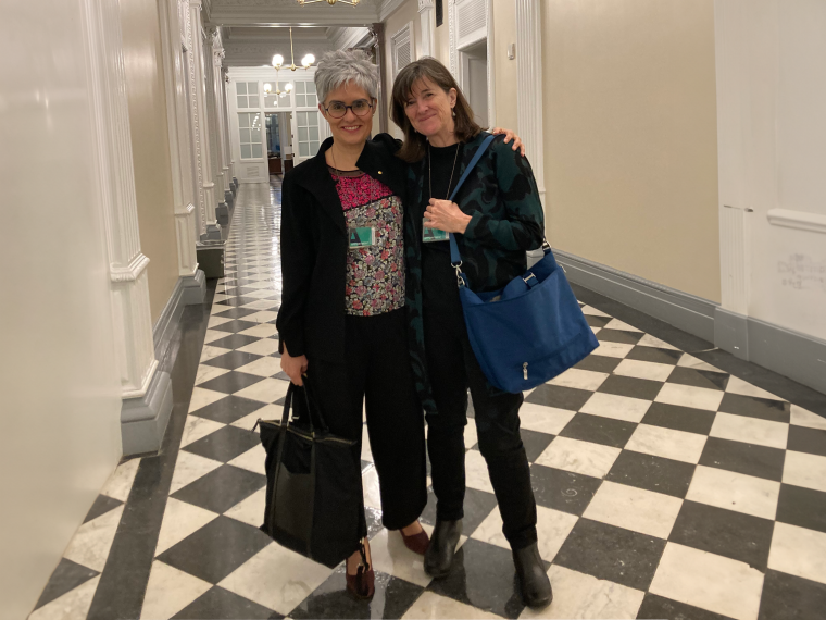 Laura López-Hoffman poses with ennifer Montoya, BLM, with Laura in the Eisenhower Executive Office Building (White House complex) following a CEQ reception. The photo was taken in a long, white hallway with black-and-white checkered tile floors.
