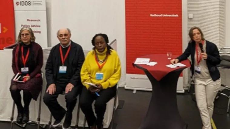 Panelists sit in tall chairs on the left as the moderator leans on a high-top table on the right side of the screen. A red and white IDOS banner stands upright behind the speakers in the top left corner of the image.