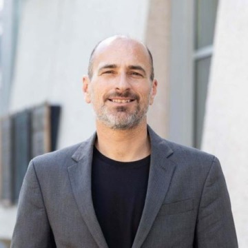 A portrait of Professor Sergio Puig in a black t-shirt and gray blazer with a white building in the background.
