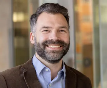 A portrait of Professor Jonathan Bean, College of Architecture, Planning and Landscape Architecture, in a brown jacket and light blue shirt