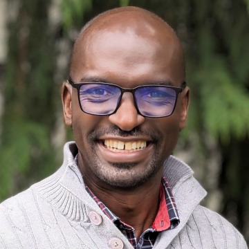 Headshot of Gordon Okumu in a gray collared sweater and plaid button-up shirt outdoors in front of greenery.