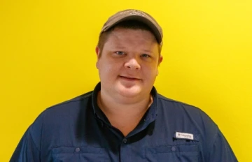 Corey Callaghan poses in a dark blue shirt and brown baseball cap in front of a yellow background.