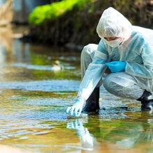 A person in full body PPE kneels in a stream to collect water in a test tube